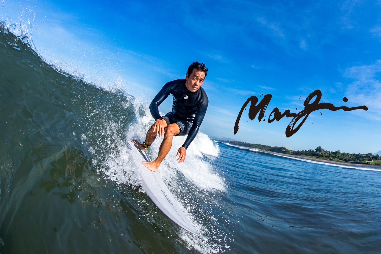 image1 - SHOP NEWS | ムラサキスポーツオンラインストア 通販 Surfer wearing a black wetsuit rides a curling ocean wave with a distant coastline under a bright blue sky.
