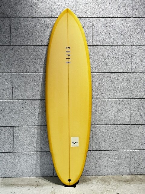 s-IMG_3504 - SHOP NEWS | ムラサキスポーツオンラインストア 通販 Yellow surfboard leaning against a gray brick wall; a small label is near the tail tip and the board is upright