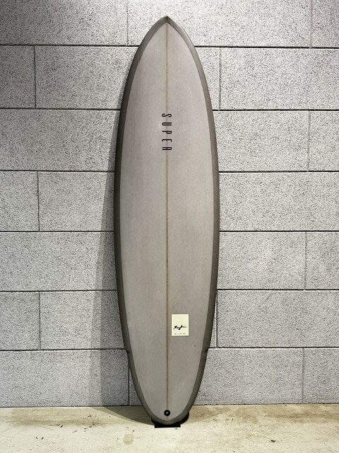 s-IMG_3506 - SHOP NEWS | ムラサキスポーツオンラインストア 通販 Gray surfboard standing upright against a gray brick wall, with a small sticker near the bottom center and a fin slot at the base edge.