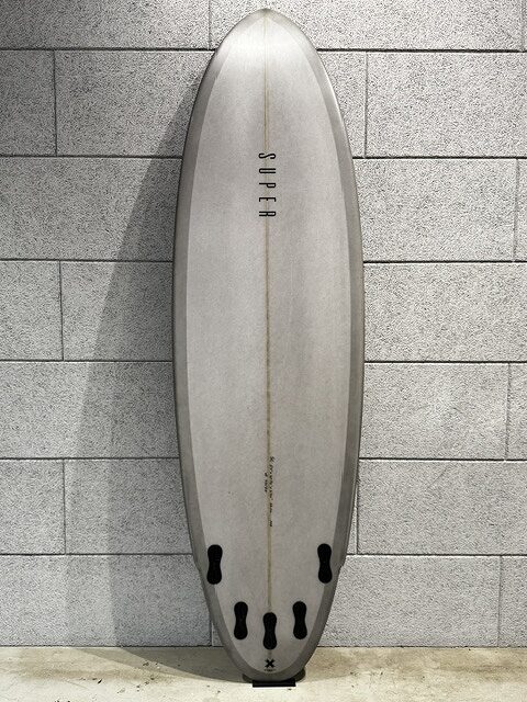 s-IMG_3507 - SHOP NEWS | ムラサキスポーツオンラインストア 通販 Light gray surfboard with a central stringer and 'SUPER' printed vertically, standing against a gray tile wall; tail shows four black fin boxes.