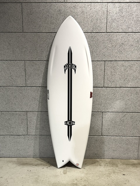 White surfboard with a bold black vertical stripe and logos, leaning against a gray brick wall.