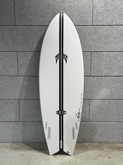 White surfboard with a bold black vertical stripe runs down the center, propped against a gray tiled wall and showing fin box slots at the bottom left and right.