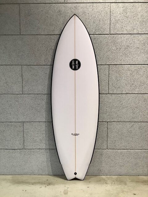 s-IMG_6653 - SHOP NEWS | ムラサキスポーツオンラインストア 通販 White surfboard with a brown center stringer leans against a gray cinder block wall, featuring a black circular logo midboard.