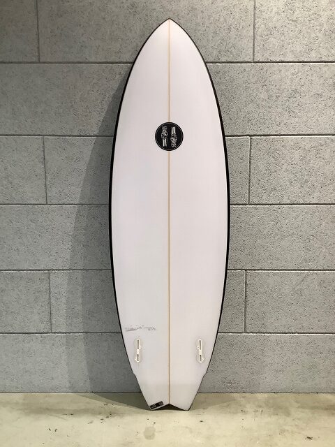 s-IMG_6654 - SHOP NEWS | ムラサキスポーツオンラインストア 通販 White surfboard with beige stringer, leaning against a gray brick wall, showing two small fin boxes near the tail and black rails.