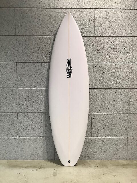 s-IMG_6655 - SHOP NEWS | ムラサキスポーツオンラインストア 通販 White surfboard standing upright against a gray cinder block wall in an indoor setting.