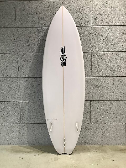 s-IMG_6656 - SHOP NEWS | ムラサキスポーツオンラインストア 通販 White surfboard standing upright against a gray brick wall, with a vertical stringer and a black logo midboard; two small fins near the tail visible.