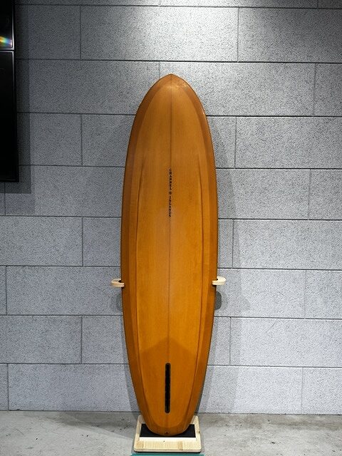 Wooden surfboard standing upright on a small display stand against a gray tiled wall.