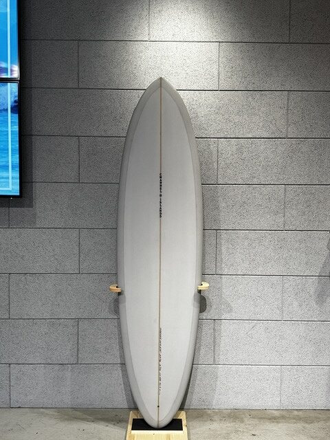 White surfboard standing upright on a small wooden rack against a gray tiled wall, ready for display or storage.