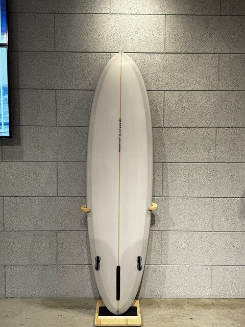 White surfboard standing upright on a wooden stand against a gray tiled wall indoors, showing a center stringer.