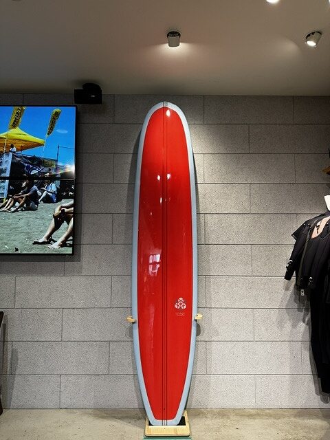Red surfboard on a wooden stand, standing upright against a gray brick wall inside a shop, with a beach photo display to the left.