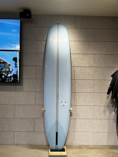 Light-blue surfboard standing upright on a wooden rack against a gray brick wall in a shop showroom.