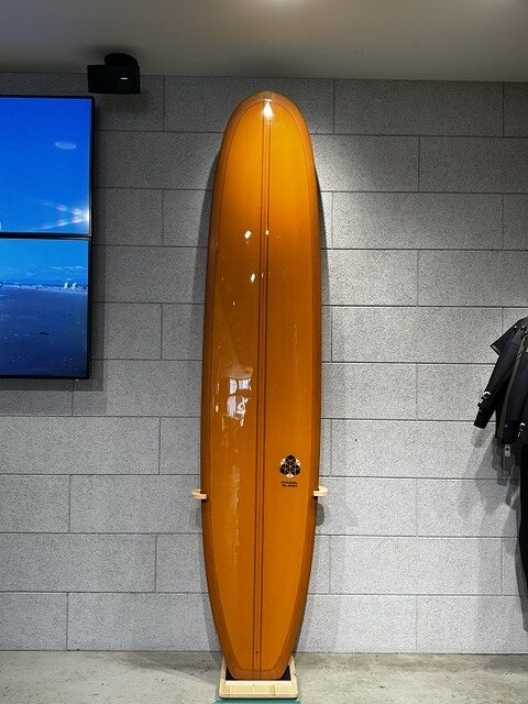 Orange longboard surfboard on a display stand against a gray tiled wall in a shop/surf shop setting.