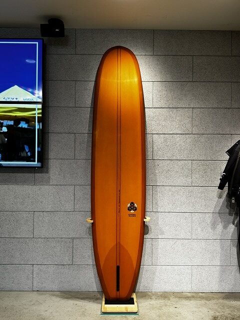 Tall orange surfboard standing upright on a small wooden stand against a gray tiled wall in an indoor showroom, with a screen to the left and a jacket to the right.