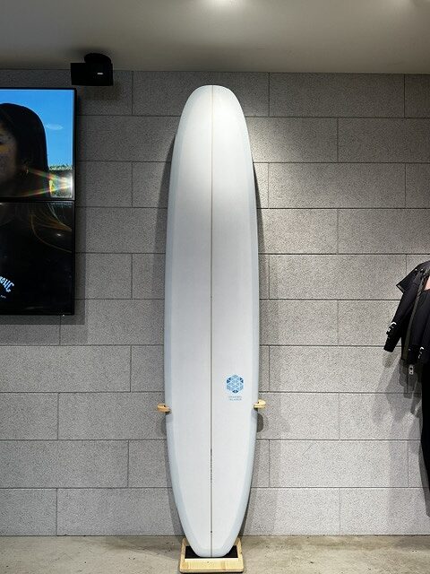 White surfboard standing upright on a small wooden rack against a gray tiled wall in a showroom/store setting.