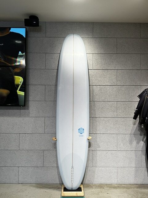 Tall white surfboard standing upright on a wooden stand against a gray brick wall, with a small logo near the tail.