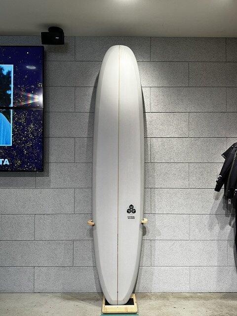 White surfboard standing upright on a wooden stand against a gray tiled wall, logo near the bottom, indoors with a wetsuit on the right and a display screen on the left.