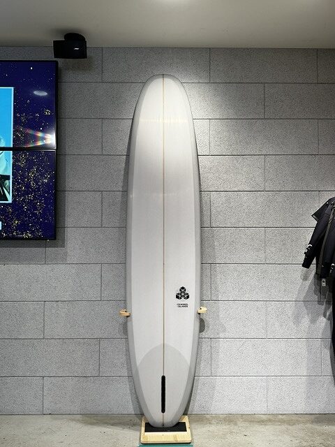 White surfboard standing upright against a gray tiled wall on a wooden stand inside a showroom, with a small logo near the center.