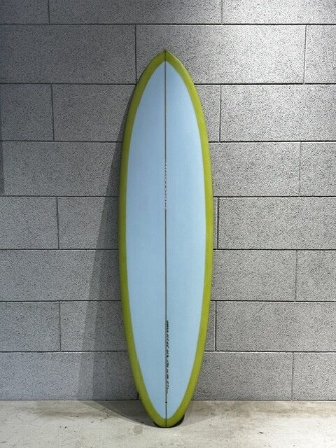 Vertical surfboard with pale blue center and olive-green edge propped against a gray tile wall in a showroom setting.