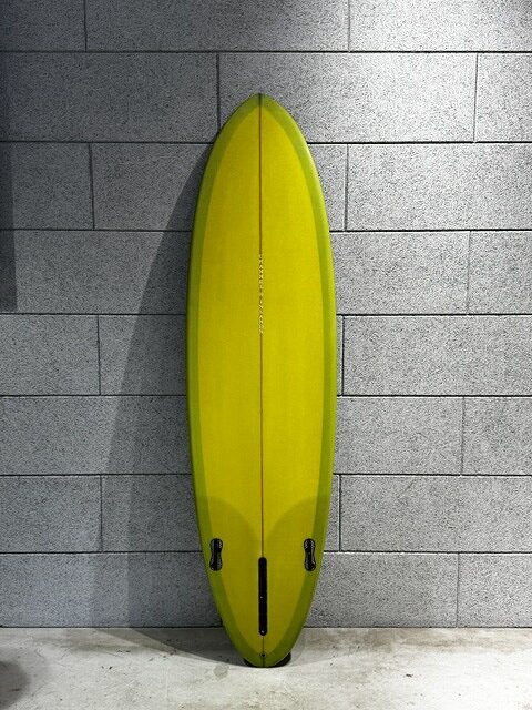 Yellow surfboard standing upright against a gray brick wall, with two fin boxes at the bottom and a central channel in the board surface.
