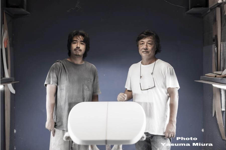 Two men stand in a surfboard shaping shop beside a white surfboard blank, posing for the photo near a blue wall panelled workshop area with tools visible on the sides.