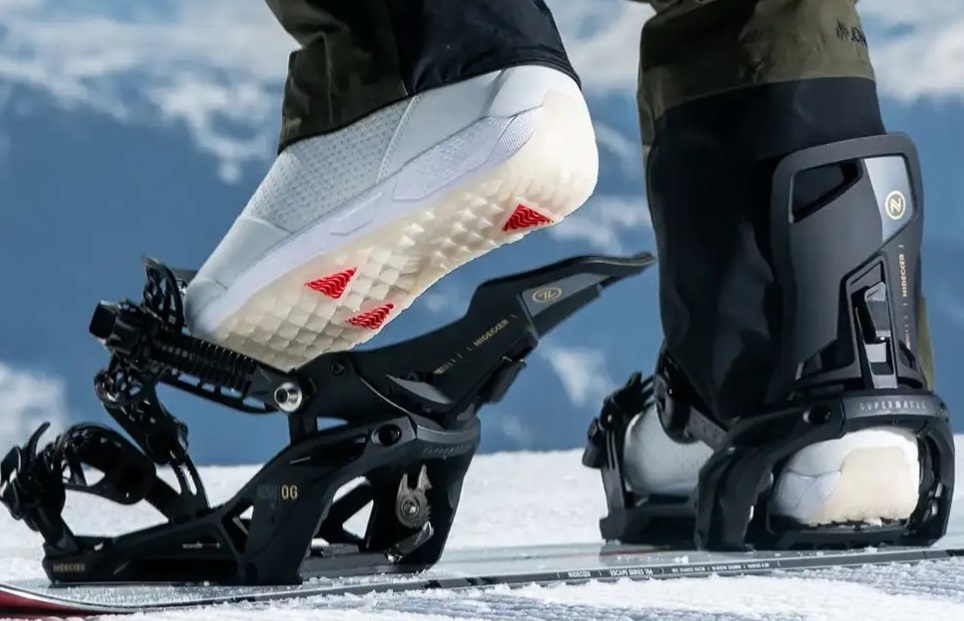 Close-up of a snowboard boot stepping into a black binding on a snowy slope with mountains in the background.