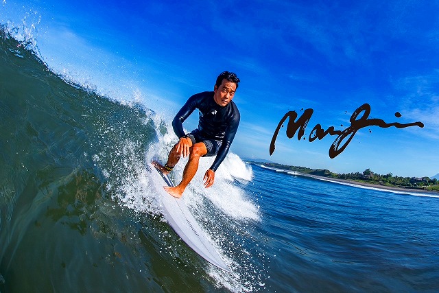 マージン 11 - SHOP NEWS | ムラサキスポーツオンラインストア 通販 Surfer in a black wetsuit rides a curling green wave on a white board under a bright blue sky, coastline in the distance.