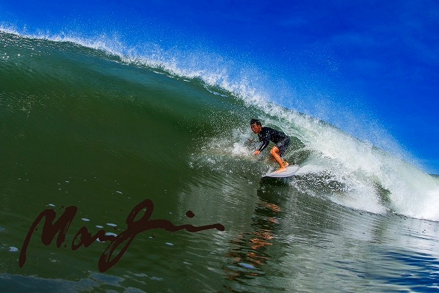 マージン 22 - SHOP NEWS | ムラサキスポーツオンラインストア 通販 Surfer riding a green curling wave under a clear blue sky; watermark signature in the lower-left corner.
