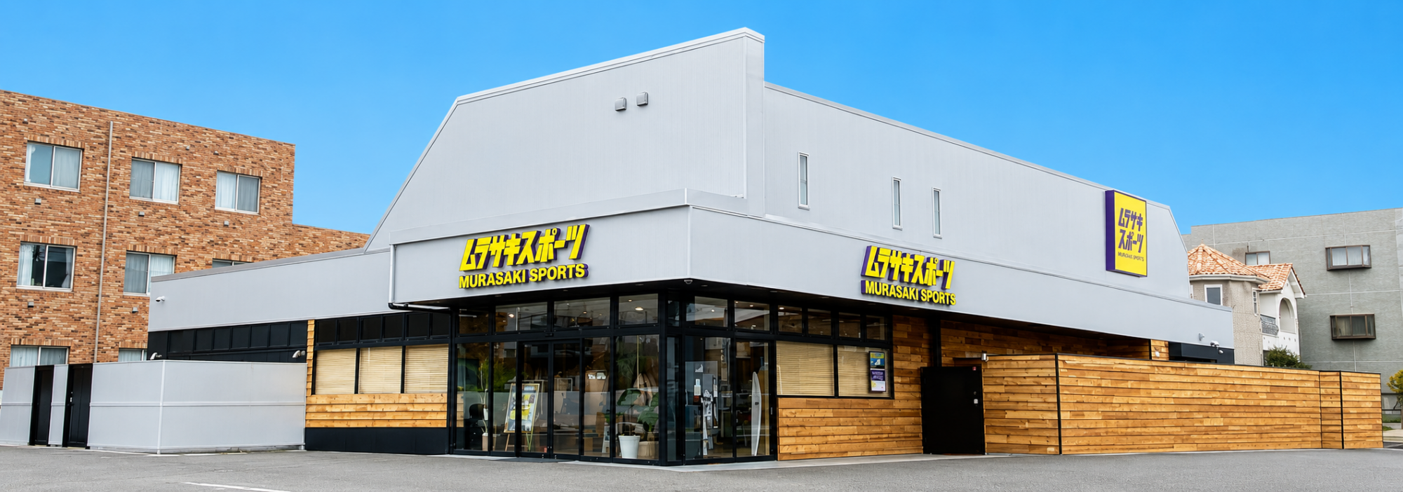 Murasaki Sports storefront with large glass windows, gray facade, and wooden fencing, under a clear blue sky.