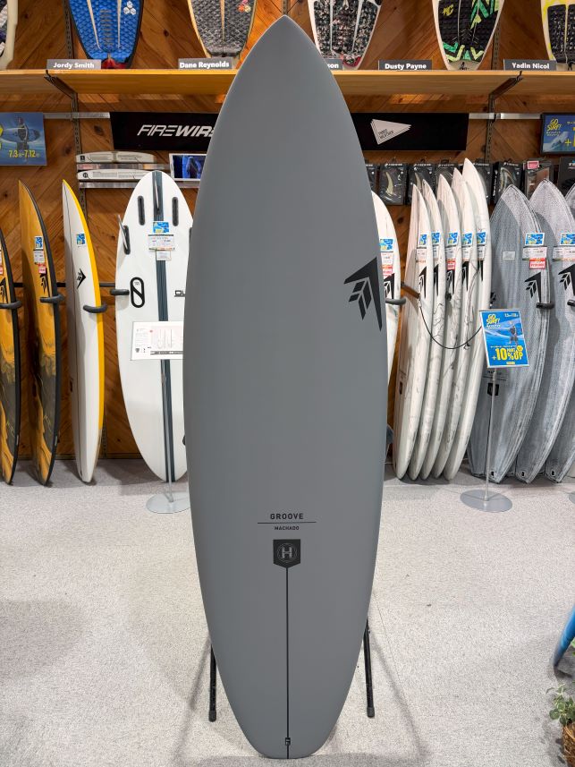 IMG_0042 - SHOP NEWS | ムラサキスポーツオンラインストア 通販 Gray surfboard on a stand in a shop, surrounded by assorted boards on shelves and racks behind it, with Groove/Machado branding visible near the bottom.