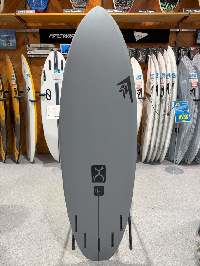 IMG_0043 - SHOP NEWS | ムラサキスポーツオンラインストア 通販 Grey surfboard on display in a shop, centered with a logo midboard and bottom fin-slot plugs, surrounded by other boards on racks and shelves beside it.