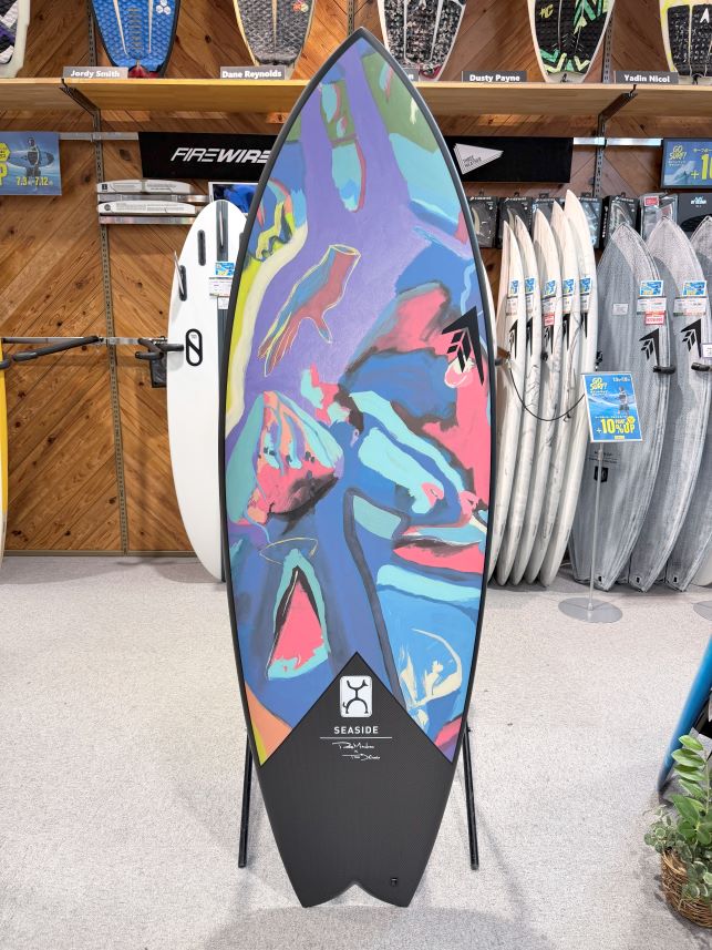 IMG_0073 - SHOP NEWS | ムラサキスポーツオンラインストア 通販 Colorful abstract-pattern surfboard on a stand in a surf shop, with other boards and racks in the background; Seaside logo near the bottom.