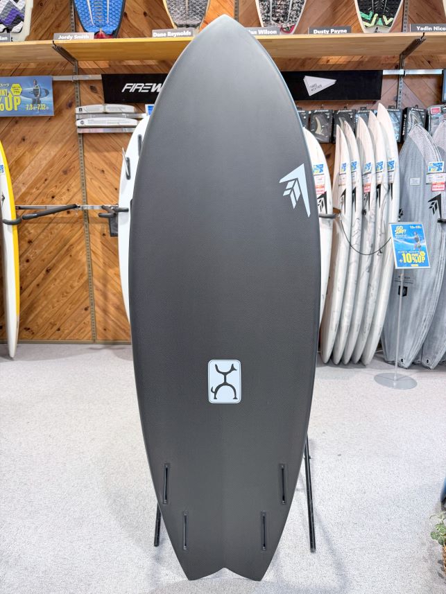 IMG_0074 - SHOP NEWS | ムラサキスポーツオンラインストア 通販 Dark gray surfboard on a display stand in a surf shop, with a small light-blue logo mid‑board and other boards visible in the background.