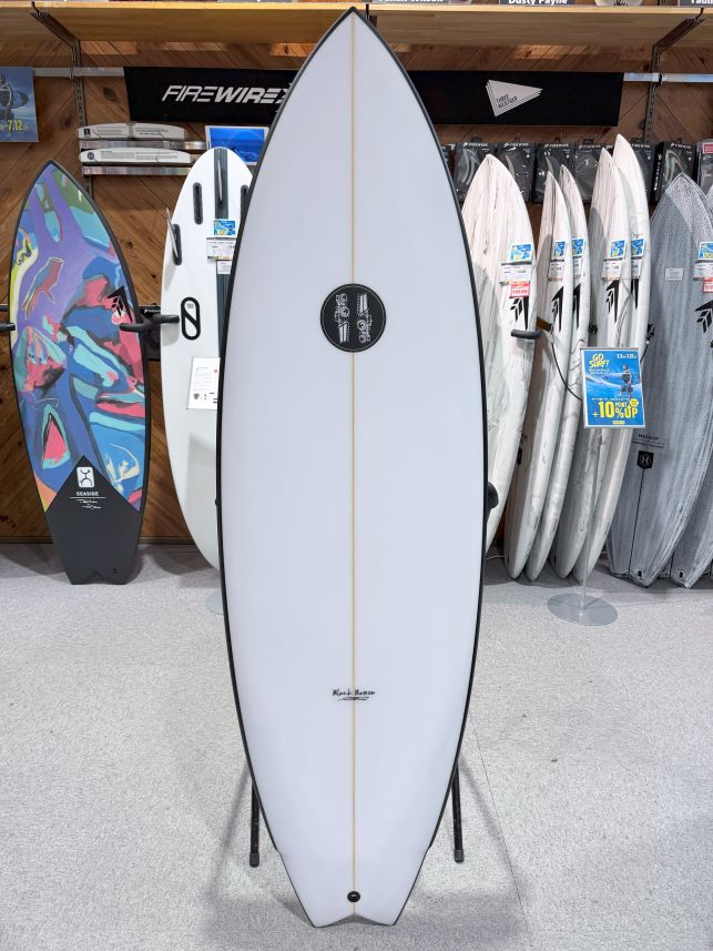 White surfboard on a stand in a surf shop, centered with other boards and racks in the background.