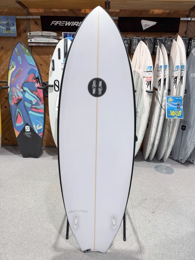 White surfboard on a display stand inside a surf shop, with other boards and gear visible in the background.