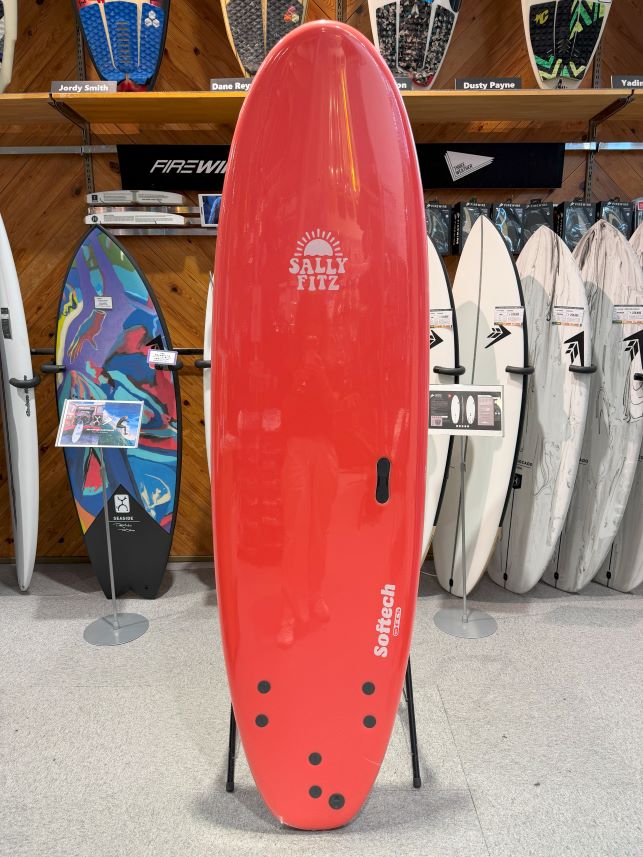 Red surfboard on display in a shop, featuring the Sally Fitz branding and Softtech logo with other boards in the background on shelves and racks.