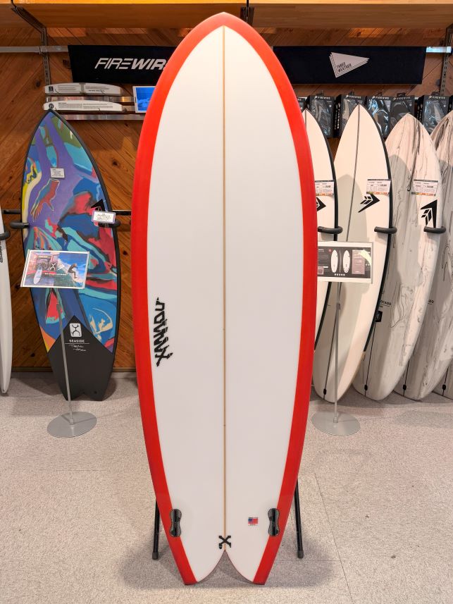 IMG_0170 - SHOP NEWS | ムラサキスポーツオンラインストア 通販 White surfboard with red rails on a display stand in a surf shop, with other boards in the background.
