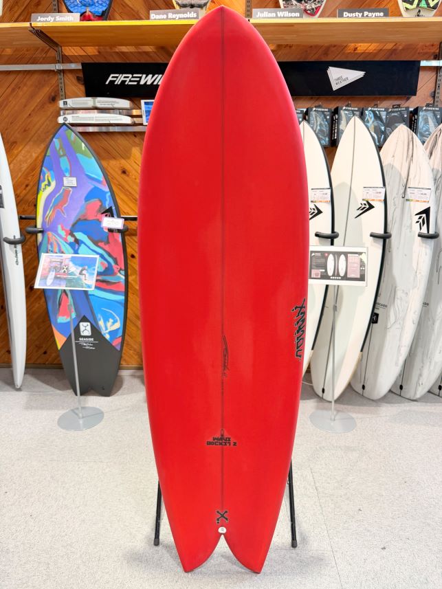 IMG_0173 - SHOP NEWS | ムラサキスポーツオンラインストア 通販 Red surfboard displayed upright on a stand inside a shop, with other boards and racks visible in the background.