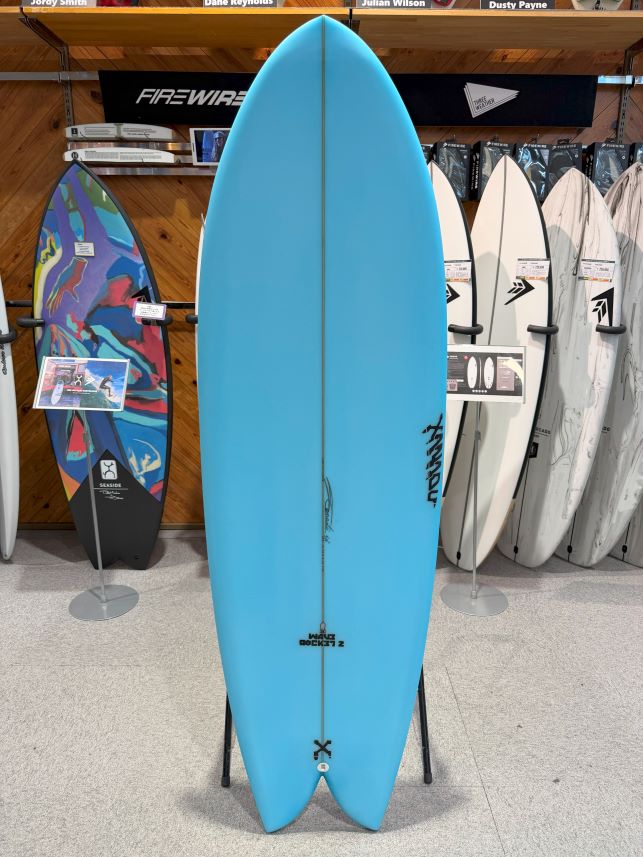 IMG_0175 - SHOP NEWS | ムラサキスポーツオンラインストア 通販 Bright blue surfboard on a display stand in a surf shop, with other boards visible in the background.