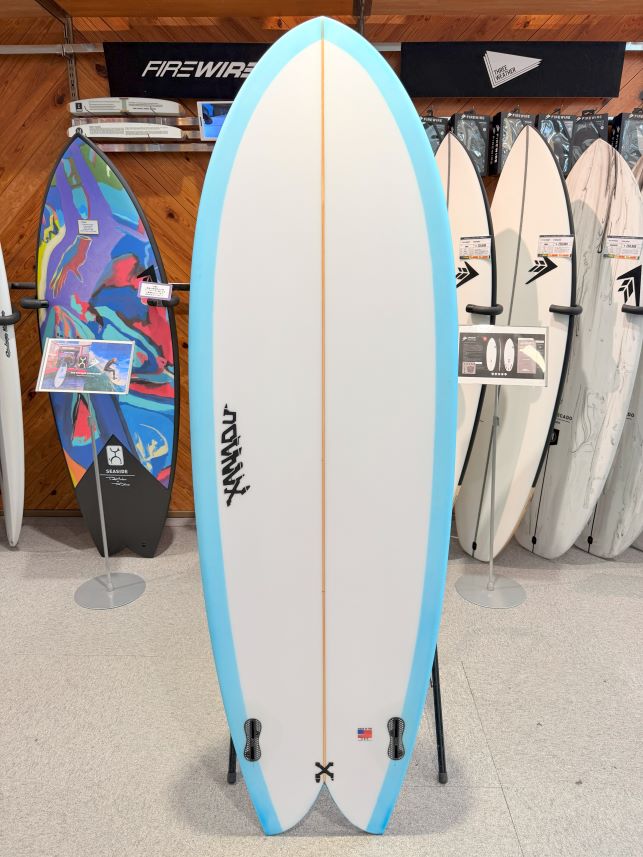 IMG_0177 - SHOP NEWS | ムラサキスポーツオンラインストア 通販 White surfboard with a light blue edge on a display rack in a surf shop, with colorful boards and signs in the background.