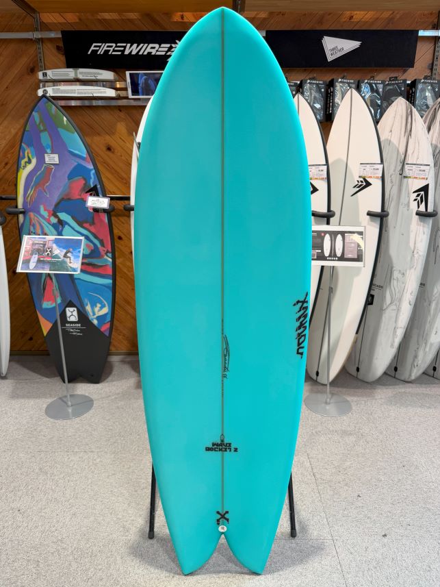 IMG_0179 - SHOP NEWS | ムラサキスポーツオンラインストア 通販 Turquoise surfboard on a display stand in a surf shop, flanked by colorful boards in the background.