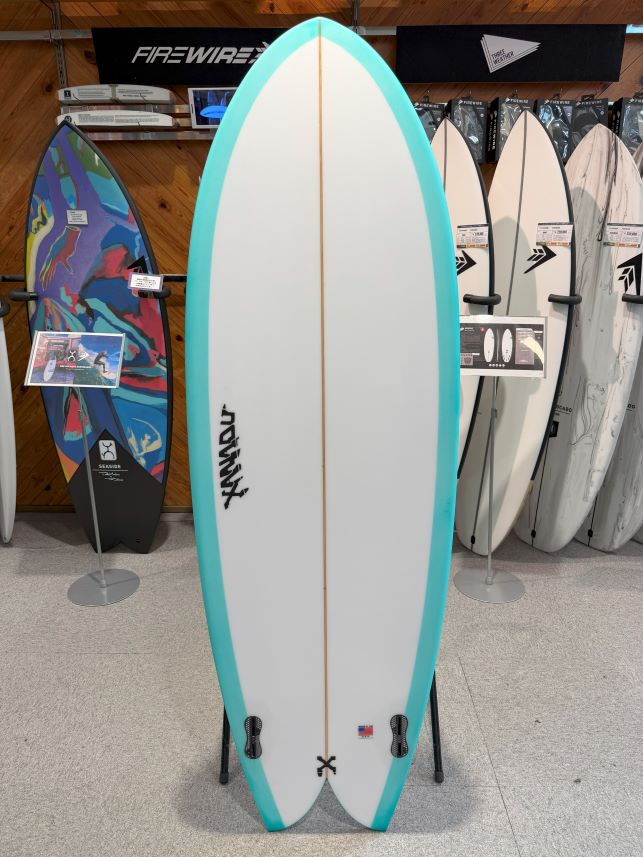 IMG_0182 - SHOP NEWS | ムラサキスポーツオンラインストア 通販 White surfboard with turquoise rails on a showroom display, a tan stringer down the center, and small black branding near the tail, with other boards in the background.