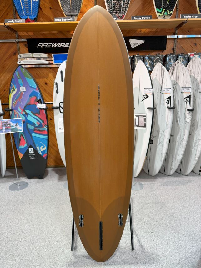 IMG_0222 - SHOP NEWS | ムラサキスポーツオンラインストア 通販 Tall brown surfboard on a display stand in a surf shop, surrounded by other boards on walls and racks, center stage.