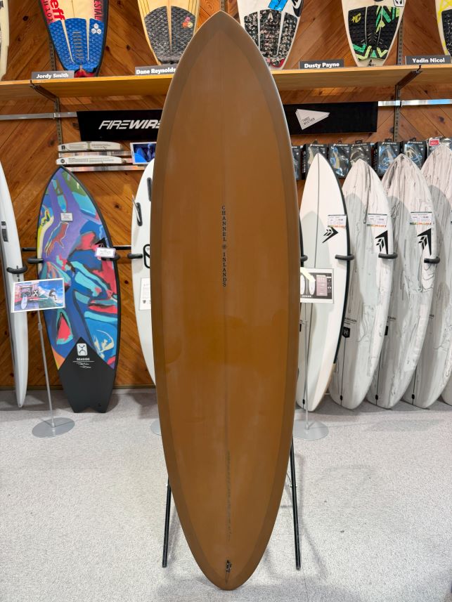 IMG_0223 - SHOP NEWS | ムラサキスポーツオンラインストア 通販 Brown longboard on a display stand in a surf shop, with colorful boards lined up on the wall behind it.