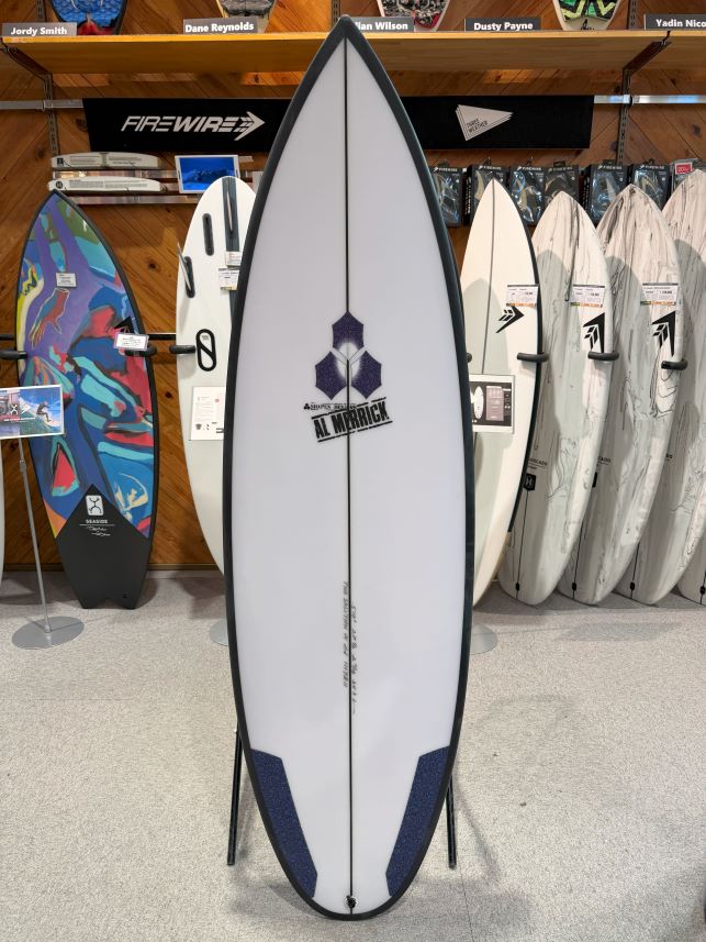 IMG_0237 - SHOP NEWS | ムラサキスポーツオンラインストア 通販 White surfboard with black rails and blue tail pads on a stand, featuring the Al Merrick logo, displayed in a shop.