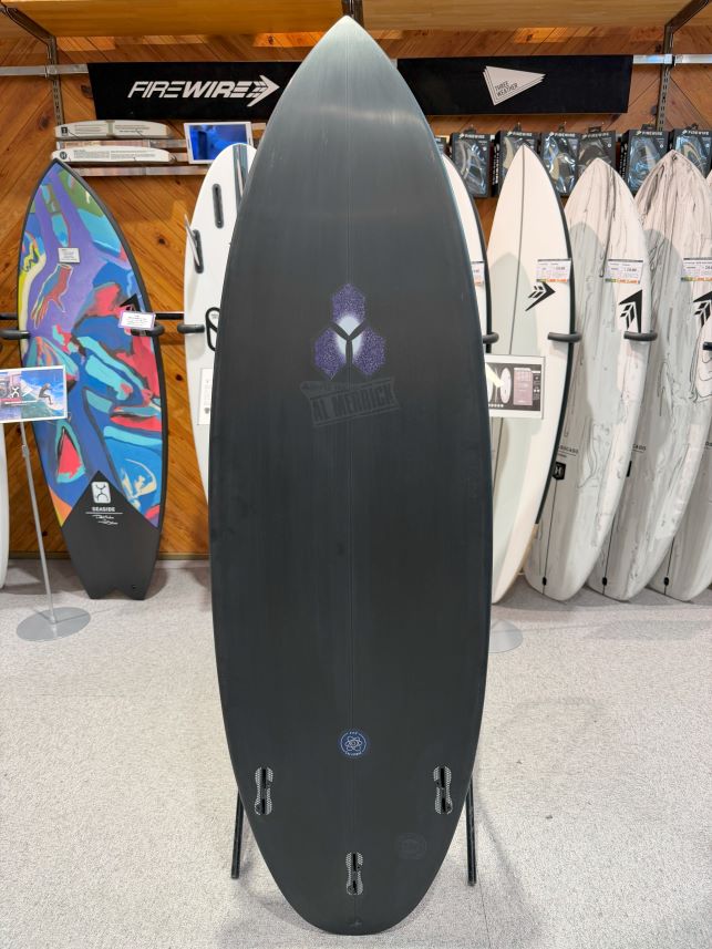 IMG_0238 - SHOP NEWS | ムラサキスポーツオンラインストア 通販 Black surfboard on a stand in a surf shop, with a purple geometric logo and multiple leash plugs near the tail, surrounded by other boards on racks.