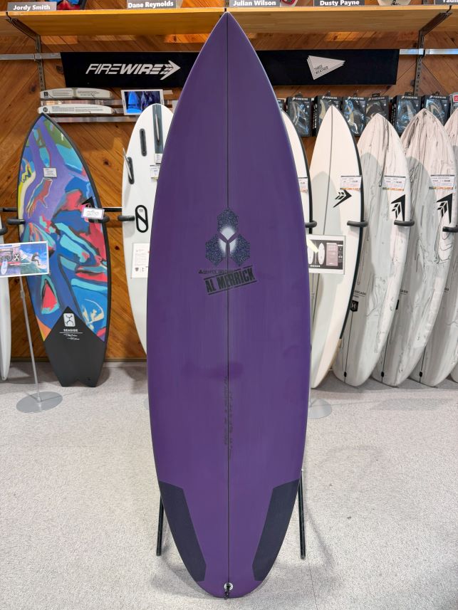IMG_0240 - SHOP NEWS | ムラサキスポーツオンラインストア 通販 Purple surfboard on a stand in a showroom, with a central logo and dark tail patches visible among other boards in the shop.