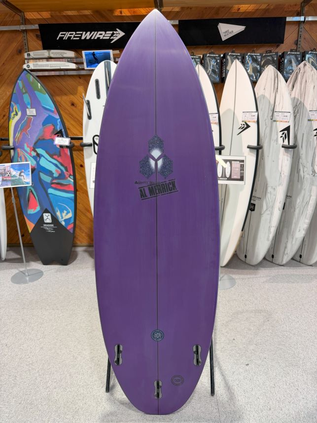 IMG_0241 - SHOP NEWS | ムラサキスポーツオンラインストア 通販 Purple surfboard on a display rack in a store, flanked by other boards and brand banners; features multiple fin boxes on the bottom and a central logo/stencil near the middle.