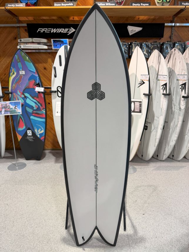 IMG_0243 - SHOP NEWS | ムラサキスポーツオンラインストア 通販 Center gray surfboard on display rack in a shop, with other boards and a wood-paneled wall in the background.