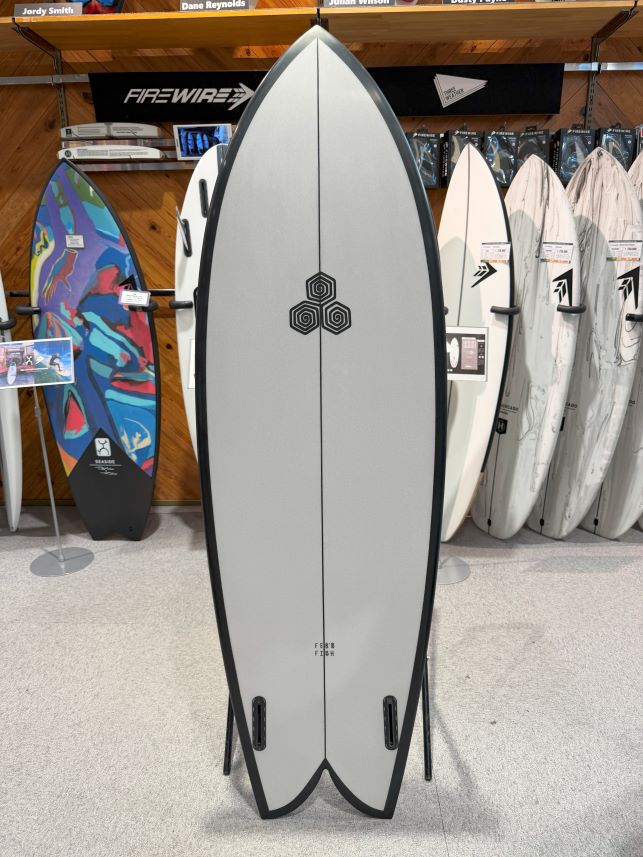 IMG_0244 - SHOP NEWS | ムラサキスポーツオンラインストア 通販 Gray surfboard on a stand in a shop, centerboard with a geometric logo and other boards in the background.
