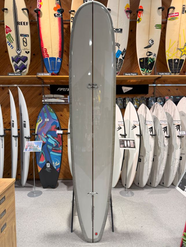 Tall gray surfboard on a stand displayed in a surf shop, with a row of boards and colorful stickers on the wall behind it.